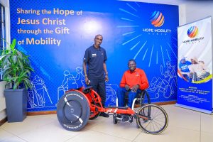 Eliud Kipchoge standing next to Michael Panther in his wheelchair alongside 'The Red Ferrari' racing wheelchair at Hope Mobility Kenya.